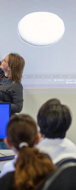 A lecturer pointing at a 3D design projected on a screen while students follow along on their computers in a CAD classroom. A lecturer pointing at a 3D design projected on a screen while students follow along on their computers in a CAD classroom.